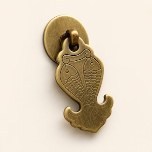 May include: A brass-coloured metal furniture pull featuring a circular backplate and a drop handle shaped like a stylised fish motif. The handle displays an engraved design of two fish facing each other with detailed scale patterns and a flared tail base.