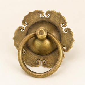 Puede incluir: Un tirador de mueble de latón con un anillo circular unido a una placa decorativa. La placa tiene un borde festoneado en forma de nube con intrincados detalles de puntos a lo largo del perímetro. El metal tiene un acabado dorado antiguo cepillado.