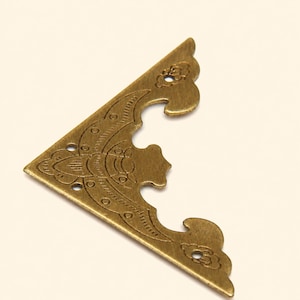 Può includere: Un paraspigoli in metallo color ottone di forma triangolare, caratterizzato da un intricato motivo inciso con bordi curvi e tre piccoli fori circolari per il fissaggio. La superficie presenta una finitura spazzolata e anticata.