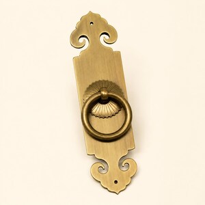 May include: A vertical brass door pull featuring a rectangular backplate with ornate, scrolled ends and a central circular ring handle attached to a sunburst-patterned mounting plate. The metal has a brushed finish with visible screw holes.