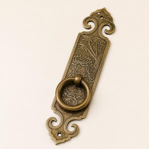 May include: A brass-toned metal door pull featuring an elongated rectangular backplate with ornate scrollwork at the top and bottom. The surface is embossed with detailed bamboo and floral motifs, and a circular ring handle is attached to the center.