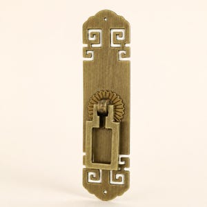 Peut inclure: Une poignée de meuble rectangulaire en laiton avec une finition brossée, dotée d'une poignée tombante ornée fixée à une plaque circulaire en forme de soleil. La plaque arrière allongée présente des découpes géométriques complexes.
