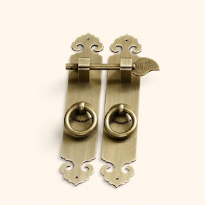 May include: A pair of antique brass door pulls featuring elongated rectangular plates with decorative scalloped ends. Each plate includes a circular ring handle and a central mounting hole. A horizontal sliding latch bar with a leaf-shaped end.