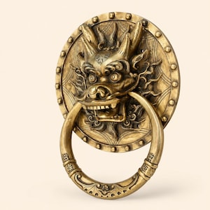 May include: A circular brass door knocker featuring a detailed three-dimensional dragon head sculpture at the center. The dragon has prominent horns, wide eyes, and an open mouth holding a large, smooth, circular ring with engraved markings.