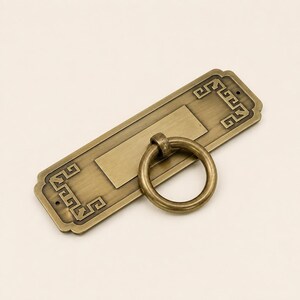 May include: A rectangular brass drawer pull featuring a central circular ring handle attached to a flat backplate. The backplate has a brushed metallic finish with decorative geometric Greek key patterns engraved on the left and right ends.