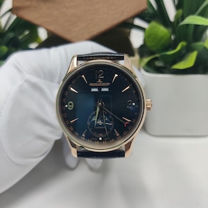 May include: A Jaeger-LeCoultre automatic wristwatch with a dark blue dial, rose gold-tone hands and indices, a black leather strap, and a moon phase display. The dial features the text JAEGER-LECOULTRE, 2030, AUG, and SWISS MADE.