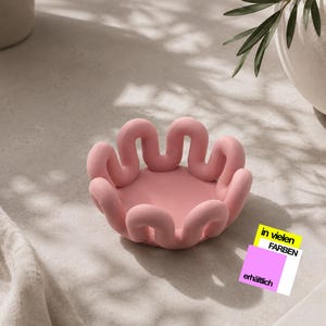Könnte beinhalten: Eine matte rosa Schale mit einem wellenförmigen, durchgehenden Rand. Neben der Schale liegt eine kleine quadratische Karte mit einem gelben Rechteck mit dem Text "in vielen FARBEN" und einem rosa Quadrat mit dem Text "erhältlich" in Schwarz.