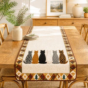 May include: A rectangular quilted table runner featuring a cream-colored center panel with six appliqued cats in various colors and patterns, bordered by a geometric diamond-shaped trim in shades of brown, green, and orange with small tree motifs.