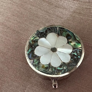May include: A round silver-toned metal pill box featuring an inlaid floral design. The centre of the lid displays a white mother-of-pearl flower with eight petals, surrounded by a dark, iridescent abalone shell inlay with green and blue tones.