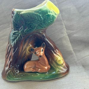 May include: A ceramic vase shaped like a hollow tree stump with a glossy finish. The exterior features moulded green leaves and brown bark textures. A small, brown, white-tailed deer figurine is positioned in the centre opening at the base.