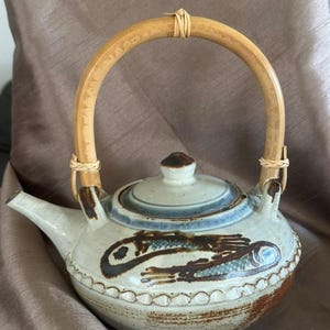 May include: A ceramic teapot with a light grey speckled glaze, featuring a hand-painted dark brown and blue fish motif on the body. The teapot has a curved bamboo handle secured with woven rattan, a matching lid, and a textured scalloped band.