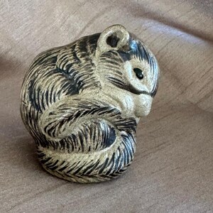 May include: A ceramic figurine of a curled-up squirrel with a textured, speckled beige finish and dark brown incised lines detailing the fur. The squirrel has a small, rounded ear, a black eye, and a compact, rounded posture with its tail tucked.
