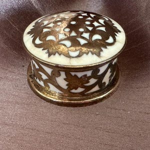 May include: A small round lidded box featuring a cream-coloured base with an intricate overlay of tarnished gold-toned metal filigree. The metalwork displays a repeating floral and leaf pattern that covers both the lid and the sides of the container.