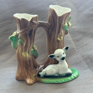 May include: A ceramic vase shaped like two brown tree trunks with green leaves and acorns, featuring a small white fawn resting at the base on a green patch. The vase has two openings at the top and a glossy finish throughout the entire structure.