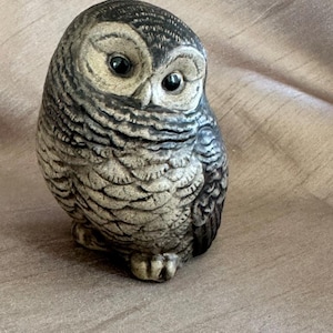 May include: A ceramic owl figurine featuring a textured, mottled grey and beige finish. The owl has large, dark, glossy eyes, a rounded head, and detailed feather patterns carved into its surface, creating a realistic and tactile appearance.