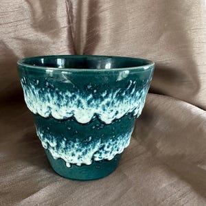 May include: A tapered ceramic planter featuring a deep teal green base colour with two horizontal bands of textured, creamy white drip glaze that create a wave-like pattern around the exterior surface of the vessel.