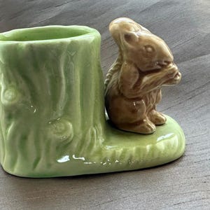 May include: A ceramic planter shaped like a light green tree stump with a textured bark surface. Attached to the side is a small, brown, glossy squirrel figurine sitting on a base, holding a nut in its paws. The piece has a smooth, glazed finish.