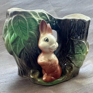 May include: A ceramic planter shaped like a hollow tree stump with textured bark and green leaves. A small, light brown rabbit with a white face and chest stands upright against the front of the stump. The interior of the planter is white.