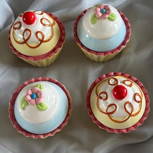 May include: Four ceramic cupcake-shaped containers with lids. Two feature yellow bases, white frosting tops with brown swirl detailing, and a red cherry accent. Two feature light blue bases, white tops, and a pink flower with green leaves.