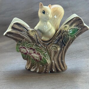 May include: A ceramic planter shaped like a tree stump with a glossy finish. A small cream-coloured squirrel sits on the centre of the stump. The stump features textured brown bark, green leaves, and small pink flowers with white centres on the sides.