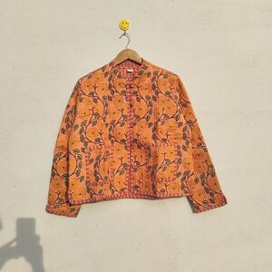 May include: A long-sleeved, quilted jacket in a vibrant orange hue, featuring a repeating pattern of dark green vines and yellow flowers. The garment includes a mandarin collar, a front button closure, and two square patch pockets on the front.