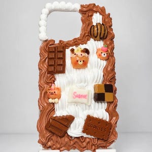 May include: A phone case decorated with white and brown whipped cream texture. It features miniature charms including a chocolate bar, a bear-shaped cupcake, a bear face, a white candy with the word Sweet, a square block, and a cookie labelled Cookies.