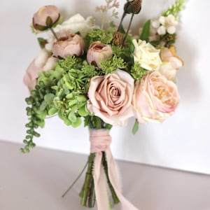 May include: A floral bouquet featuring dusty pink and cream roses, small green hydrangea clusters, and trailing green foliage. The stems are bound together with a light pink fabric ribbon that hangs down in two long, frayed-edge streamers.