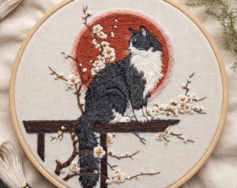 May include: A circular embroidery hoop containing a fabric panel with a hand-stitched design of a black and white cat sitting on a brown wooden branch. Behind the cat is a large, circular, rust-colored sun, surrounded by white cherry blossoms.
