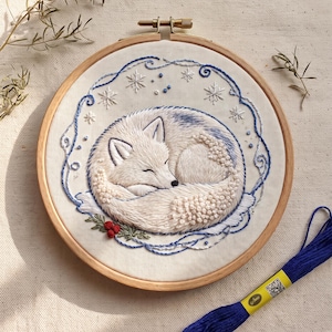 Könnte beinhalten: Ein runder Stickrahmen aus Holz mit einem weißen Stoffeinsatz, der ein handgesticktes Motiv eines zusammengerollten weißen Fuchses zeigt. Der Fuchs ist von blauen Stickereien, weißen Schneeflocken und kleinen roten Beeren umgeben.