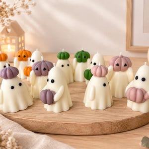 Può includere: Una collezione di candele bianche a forma di fantasma, ognuna con due occhi neri e una piccola zucca sulla testa o tra le braccia. Le zucche sono colorate di arancione, verde, viola e rosa e sono disposte su un tagliere di legno rotondo.