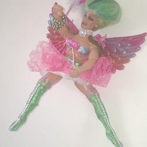 May include: A doll with green hair and a white feather, wearing pink wings, a silver bra, a pink lace skirt, and green boots with silver trim. The doll has a pearl-style choker, a beaded bracelet, and holds a small pink wand with a silver handle.