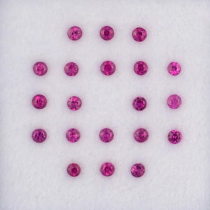 May include: Twenty-one small, round, faceted rubies in a white display case. The gems are a vibrant magenta colour and arranged in a grid pattern. The case has a white border.