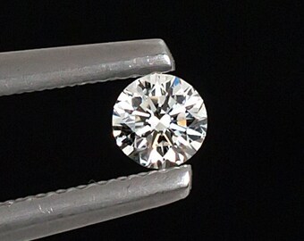 May include: A clear round diamond gemstone held in silver tweezers against a black background.
