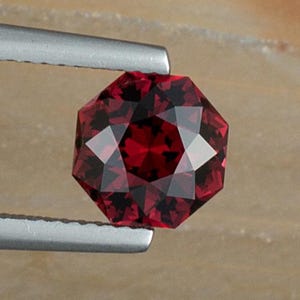 May include: A deep red gemstone with a faceted octagonal shape, held by tweezers against a brown and red background.
