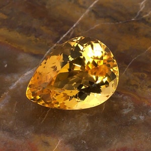 May include: A pear-shaped, faceted, golden yellow gemstone on a brown, marbled surface.