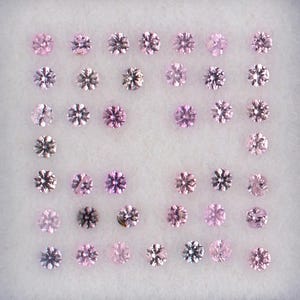 May include: A collection of small, round, pink gemstones arranged in a white tray. The gemstones have a starburst pattern and vary in shades of pink, from light to dark. The tray is set in a white frame.