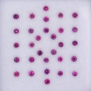 Pode incluir: Uma coleção de pequenas pedras preciosas redondas, de cor fúcsia, dispostas numa bandeja branca. As pedras preciosas têm uma aparência facetada, refletindo a luz. A bandeja está sobre um fundo branco, realçando as pedras.