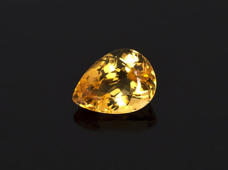 May include: A pear-shaped, faceted gemstone with a golden yellow color. The gemstone is set against a black background.