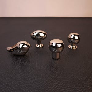 May include: Four polished silver-toned metal cabinet knobs with a reflective finish. Each knob features the engraved script text Tiffany. The set includes one bird-shaped knob, two spherical knobs, and one oval-shaped knob, all with matching stems.