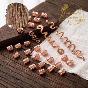 Può includere: Una collezione di accessori per capelli in oro rosa che include numerose piccole perline cilindriche con motivi intricati e due lunghe spirali. Un'immagine inserita mostra questi accessori applicati su capelli intrecciati. Il testo recita Customizable Gift Box.