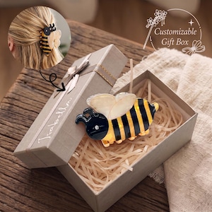 Puede incluir: Una pinza para el pelo con forma de abeja con rayas negras y amarillas, alas translúcidas de color blanco roto y un pequeño brillante transparente, presentada en una caja de regalo gris con el nombre Isabella y el texto Customizable Gift Box.