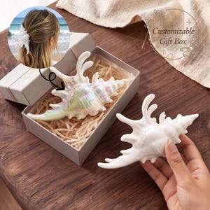 May include: An iridescent, spiky seashell hair clip with a glossy finish, presented inside a rectangular gift box filled with shredded paper filler. A second, solid white seashell hair clip is held in the foreground, showing its detailed, textured structure.