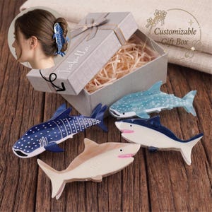 Könnte beinhalten: Eine Sammlung von vier hai-förmigen Haarspangen in Blau, Weiß und Beige mit detaillierten Mustern und Flossen. Eine Spange ist im Gebrauch zu sehen, und eine graue Geschenkbox mit dem Text Customizable Gift Box ist zur Aufbewahrung enthalten.