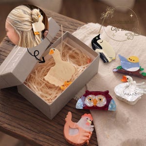 Peut inclure: Une collection de pinces à cheveux en forme d'animaux comprenant une oie, un pingouin, un oiseau bleu, un hibou, un cygne et un poulet. La pince oie est dans une boîte grise marquée Customizable Gift Box. Les pinces ont des motifs acryliques.