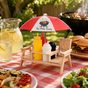 May include: A miniature wooden picnic table condiment holder featuring a red and white striped umbrella with a graphic of a grill and the text The Miller Family. It holds a yellow mustard bottle, a red ketchup bottle, and salt and pepper shakers.