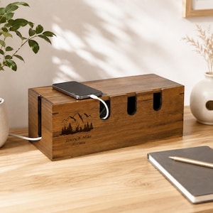 Peut inclure: Une boîte de rangement pour câbles en bois brun foncé rectangulaire avec une gravure laser représentant un paysage de montagne et de forêt, le texte Jenny & Mike et 08.18.2024. Elle possède des ouvertures pour câbles et un smartphone noir.
