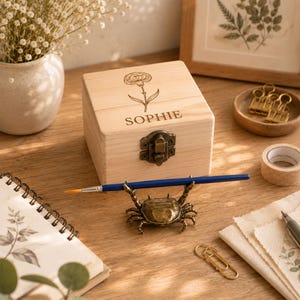 Peut inclure: Une boîte en bois clair avec un fermoir métallique à charnière, ornée d'un œillet gravé au laser et du nom SOPHIE sur le couvercle. Devant la boîte se trouve un porte-stylo en métal bronze en forme de crabe tenant un pinceau bleu.