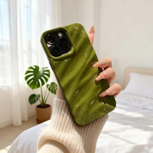 May include: An olive green phone case with a glossy, wavy, liquid-like texture. The case features a raised square camera cutout with three circular lens openings and a flash opening, designed to fit a smartphone with a triple-lens camera system.