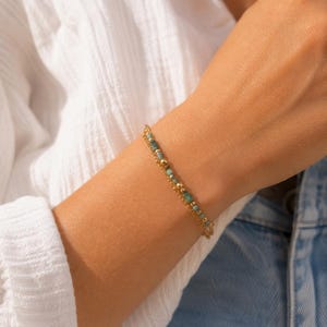 Op de afbeelding: Een verfijnde armband bestaande uit twee dunne goudkleurige kettinkjes, afgewisseld met kleine, cilindervormige turquoise kralen en piepkleine, gepolijste goudkleurige bolletjes. Het sieraad heeft een subtiel en gelaagd ontwerp.