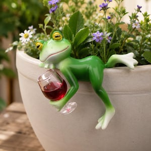 Puede incluir: Una figura de rana verde con ojos amarillos, posada en el borde de una maceta de color claro, sosteniendo un vaso transparente lleno de líquido rojo. La rana tiene un acabado liso, pies con puntas blancas y una expresión facial relajada.